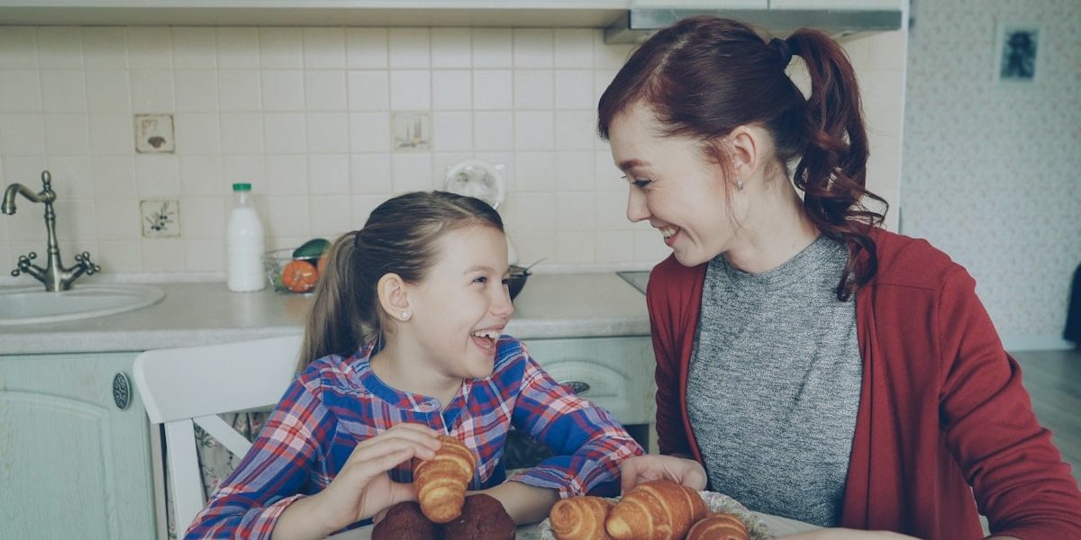 Happy young mother and cute daughter having breakfast eating muffins and talking at home in modern kitchen. Family, food, home and people concept