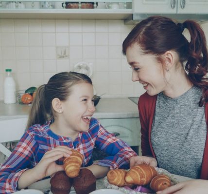 Happy young mother and cute daughter having breakfast eating muffins and talking at home in modern kitchen. Family, food, home and people concept